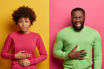Horizontal shot of dark skinned couple touch stomachs, suffer from chronic gastritis, being hungry has displeased face expressions, isolated over yellow and pink background. Healthcare concept - obrazy, fototapety, plakaty