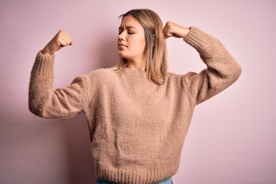 Young beautiful blonde woman wearing winter wool sweater over pink isolated background showing arms muscles smiling proud. Fitness concept.