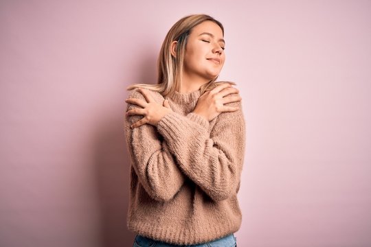 Young beautiful blonde woman wearing winter wool sweater over pink isolated background Hugging oneself happy and positive, smiling confident. Self love and self care