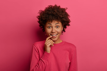 Fototapeta na wymiar Curious dark skinned woman has glad expression, keeps index finger near lips, hears interesting suggestion or information, wears crimson jumper, isolated on pink wall. Human facial expressions concept