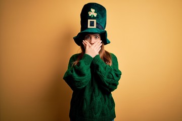 Young beautiful brunette woman wearing green hat on st patricks day celebration shocked covering...