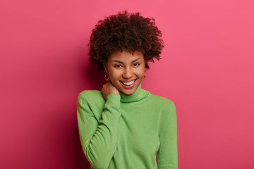 Fototapeta na wymiar Nice attractive lovely woman smiles sincerely, has bushy curly hair, touches neck, expresses pleasant emotions, hears compliment from boyfriend, isolated over pink background. Human expressions