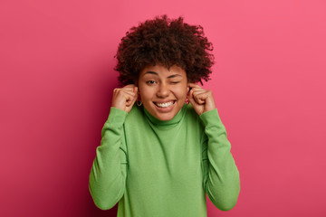 Headshot of unhappy woman with Afro hairstyle suffers from unpleasant loud noisy sound, plugs ears and winks eye, shows white teeth, dressed casually, isolated over vibrant crimson background