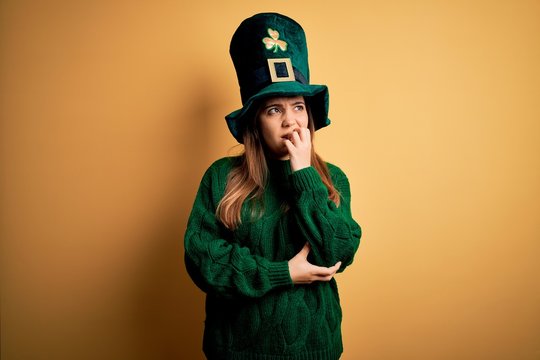 Young beautiful brunette woman wearing green hat on st patricks day celebration looking stressed and nervous with hands on mouth biting nails. Anxiety problem.