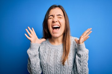 Beautiful young woman wearing casual wool sweater standing over blue isolated background celebrating mad and crazy for success with arms raised and closed eyes screaming excited. Winner concept