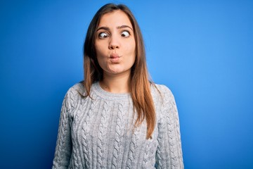 Obraz premium Beautiful young woman wearing casual wool sweater standing over blue isolated background making fish face with lips, crazy and comical gesture. Funny expression.