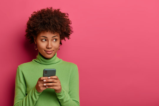Thoughtful curly hipster girl creats post on own web page, uses mobile phone, being always in touch, reads comments, concentrated aside, copy space on vibrant pink background, reads website blog