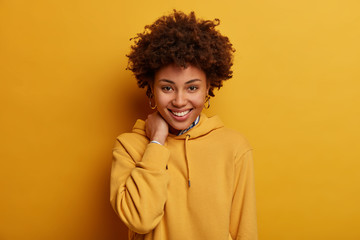 Positive teenage girl smiles happily, touches neck, wears casual hoodie, shows ideal even teeth,...