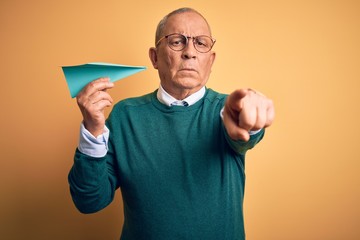 Senior handsome man holding airplane of paper standing over isolated yellow background pointing with finger to the camera and to you, hand sign, positive and confident gesture from the front