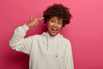 Fototapeta na wymiar Horizontal shot of glad dark skinned woman makes finger gun and shoots in temple, demonstrates suicide gesture because of boredom, wears white sweatshirt, stands in studio over rosy background