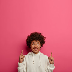 Merry smiling teenage girl has Afro haircut, points above on blank space, helps with advice, gives recommendation, shows best product in stock, recommends good shop upstairs, wears white hoodie