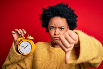 Young African American afro woman with curly hair holding vintage alarm clock with angry face,...