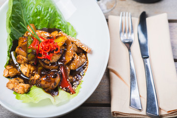 Sliced chicken fried in sweet and sour soy sauce with vegetables and herbs in a deep plate on a wooden table in a restaurant.