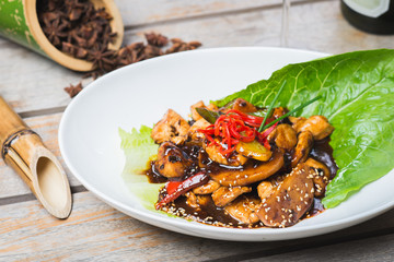 Sliced chicken fried in sweet and sour soy sauce with vegetables and herbs in a deep plate on a wooden table in a restaurant.