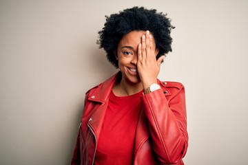 Obraz na płótnie Canvas Young beautiful African American afro woman with curly hair wearing casual red jacket covering one eye with hand, confident smile on face and surprise emotion.