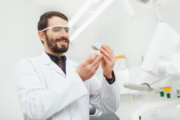 Low angle shot of a bearded handsome mature dentist smiling, examining teeth mold, working at his office, copy space
