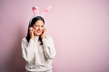 Young caucasian woman wearing cute easter rabbit ears over pink isolated background covering ears with fingers with annoyed expression for the noise of loud music. Deaf concept.