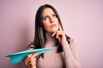 Young woman with blue eyes holding paper airplane standing over isolated pink background serious face thinking about question, very confused idea