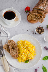 Scrambled eggs cooking from organic fresh eggs on a white plate with rye bread on a light background with a cup of coffee. Healthy breakfast. Vertical orientation. Top view, close up