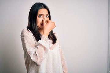 Young brunette woman with blue eyes wearing casual t-shirt over isolated white background smelling something stinky and disgusting, intolerable smell, holding breath with fingers on nose. Bad smell