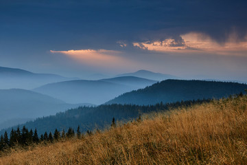 Naklejka na ściany i meble Majestic landscape of mountains at sunrise. View of the misty tops and layer hills of the mountains in the distance. Dramatic sky and rays of sunlight at morning. Concept of nature background.