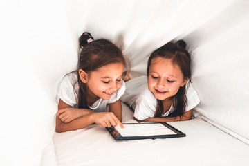 Two beautiful little sisters lying in the bed and look at the screen of a tablet, smart technology