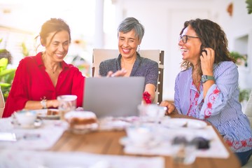 Meeting of middle age women having lunch and drinking coffee. Mature friends smiling happy using laptop at home on a sunny day