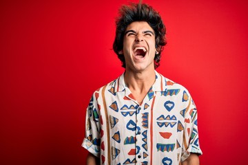 Young handsome man on vacation wearing summer shirt over isolated red background angry and mad screaming frustrated and furious, shouting with anger. Rage and aggressive concept.