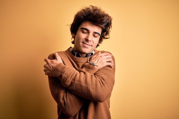 Young handsome man wearing casual shirt and sweater over isolated yellow background Hugging oneself happy and positive, smiling confident. Self love and self care