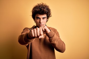 Young handsome man wearing casual shirt and sweater over isolated yellow background Punching fist to fight, aggressive and angry attack, threat and violence