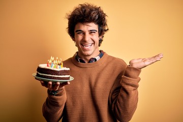 Young handsome man holding birthday cake standing over isolated yellow background very happy and excited, winner expression celebrating victory screaming with big smile and raised hands