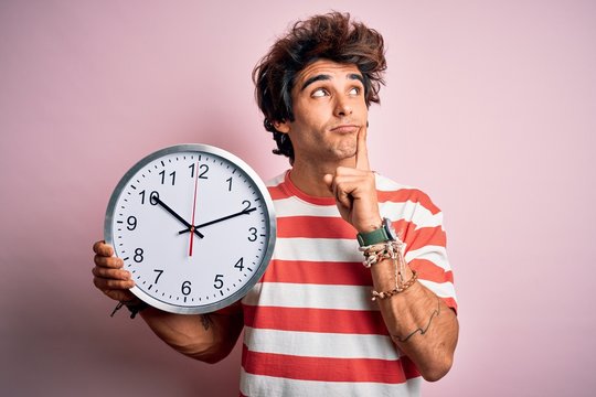 Young handsome man holding big clock standing over isolated pink background serious face thinking about question, very confused idea