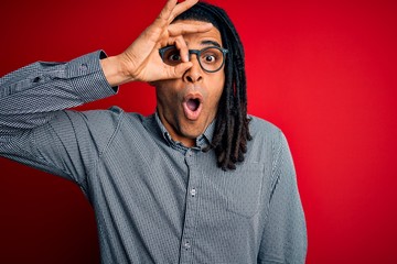 Young handsome african american man with dreadlocks wearing casual shirt and glasses doing ok gesture shocked with surprised face, eye looking through fingers. Unbelieving expression.