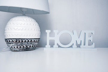 Bedside ceramic lamp with textile lampshade on the white table with the sign home, cozy home concept in grey pastel colors