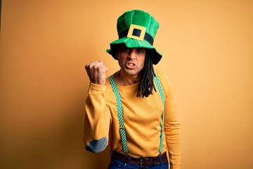 Young african american man wearing green hat celebrating saint patricks day angry and mad raising fist frustrated and furious while shouting with anger. Rage and aggressive concept.