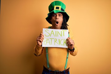 Young african american man wearing green hat holding banner celebrating saint patricks day scared...