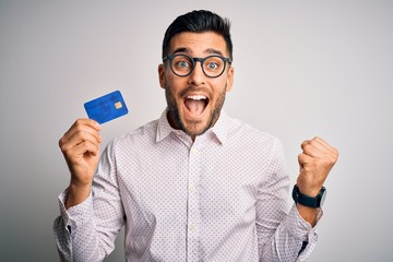 Young business man holding credit card over isolated background screaming proud and celebrating...