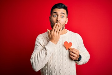 Young handsome man holding red shape heart as romantic and health symbol cover mouth with hand shocked with shame for mistake, expression of fear, scared in silence, secret concept
