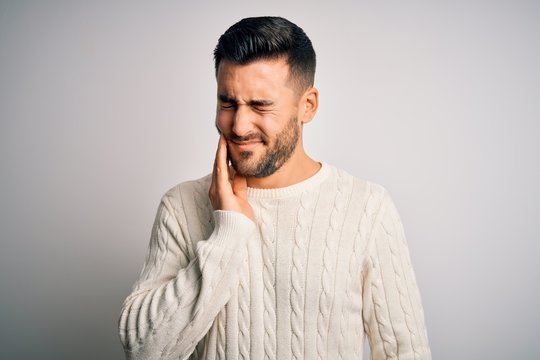 Young handsome man wearing casual sweater standing over isolated white background touching mouth with hand with painful expression because of toothache or dental illness on teeth. Dentist
