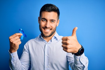Young handsome man holding blue ribbon as prostate campaing support over blue background happy with...