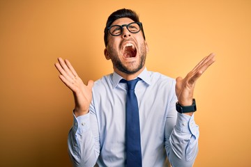 Young handsome businessman wearing tie and glasses standing over yellow background celebrating mad and crazy for success with arms raised and closed eyes screaming excited. Winner concept