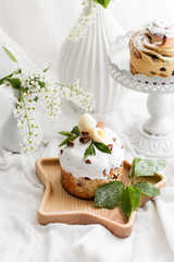 Kulich Traditional Easter sweet bread decorated with green leaves and a white egg, a candy on a wooden stand on a white fabric background. round vase with white spring flowers.Copy space.