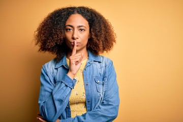 Young african american woman with afro hair wearing casual denim shirt over yellow background asking to be quiet with finger on lips. Silence and secret concept.