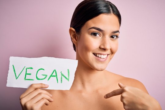 Young beautiful girl holding paper with vegan message over isolated pink background with surprise face pointing finger to himself