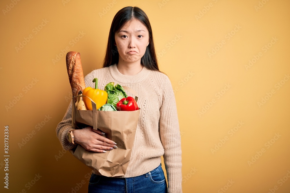 Wall mural young asian woman holding paper bag of fresh healthy groceries over yellow isolated background depre