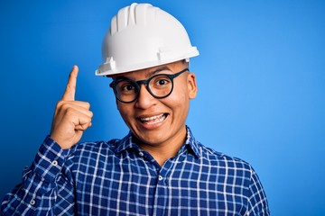 Young handsome engineer latin man wearing safety helmet over isolated blue background pointing finger up with successful idea. Exited and happy. Number one.