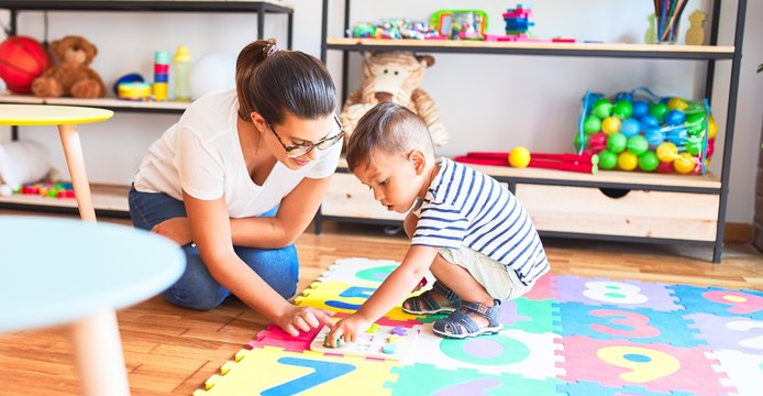 Beautiful teacher and toddler boy sitting on puzzle playing with numbers at kindergarten