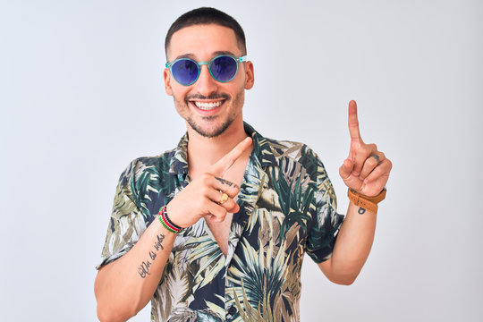 Young handsome man wearing Hawaiian sumer shirt and blue sunglasses over isolated background smiling and looking at the camera pointing with two hands and fingers to the side.
