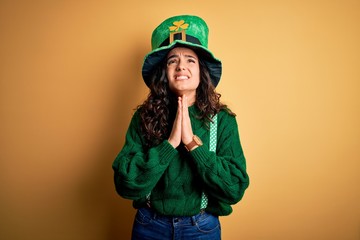 Beautiful curly hair woman wearing green hat with clover celebrating saint patricks day begging and praying with hands together with hope expression on face very emotional and worried. Begging.