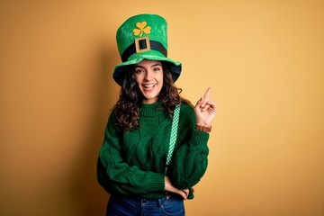 Beautiful curly hair woman wearing green hat with clover celebrating saint patricks day with a big smile on face, pointing with hand and finger to the side looking at the camera.